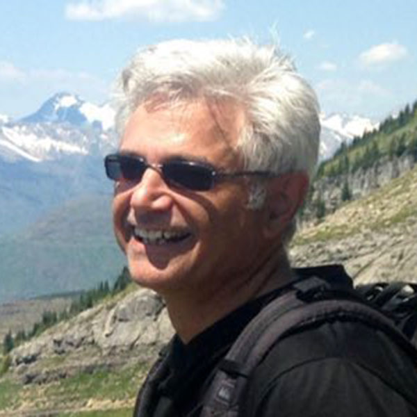 A headshot of Byron Santangelo wearing a backpack while hiking in the mountains.