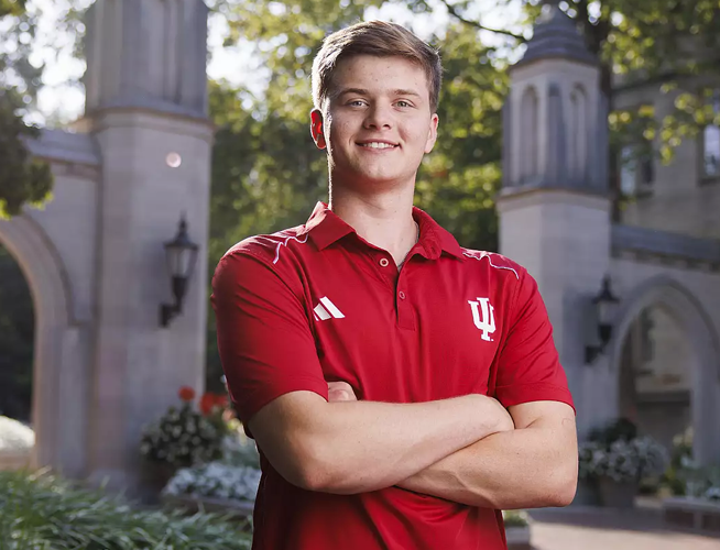 A smiling male IU student wearing a crimson IU polo shirt and standing in front of the Sample Gates.