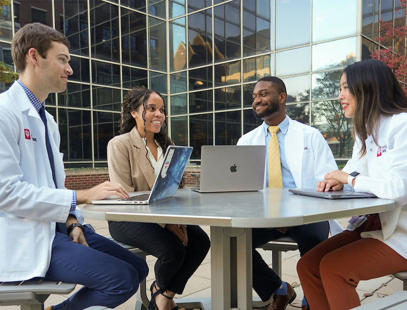 Four medical students work together on laptops in an outdoor space.