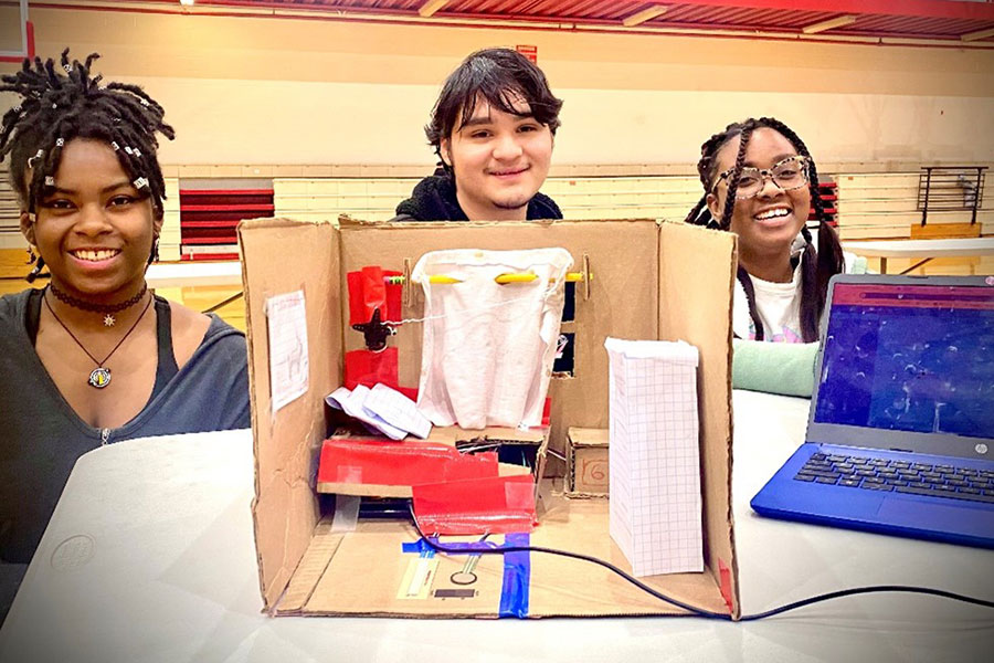 3 students stand behind a diorama connected to a laptop.