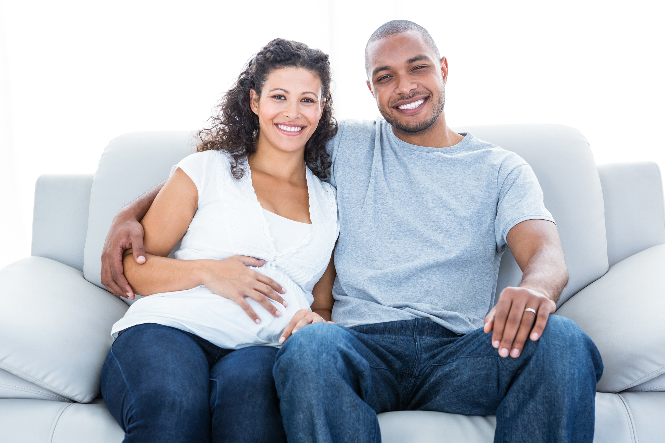 Cheerful multiethnic couple expecting a baby sitting on sofa at home.