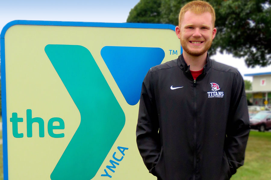 A male student wearing a Titans Cross Country jacket stands in front of an exterior YMCA sign.