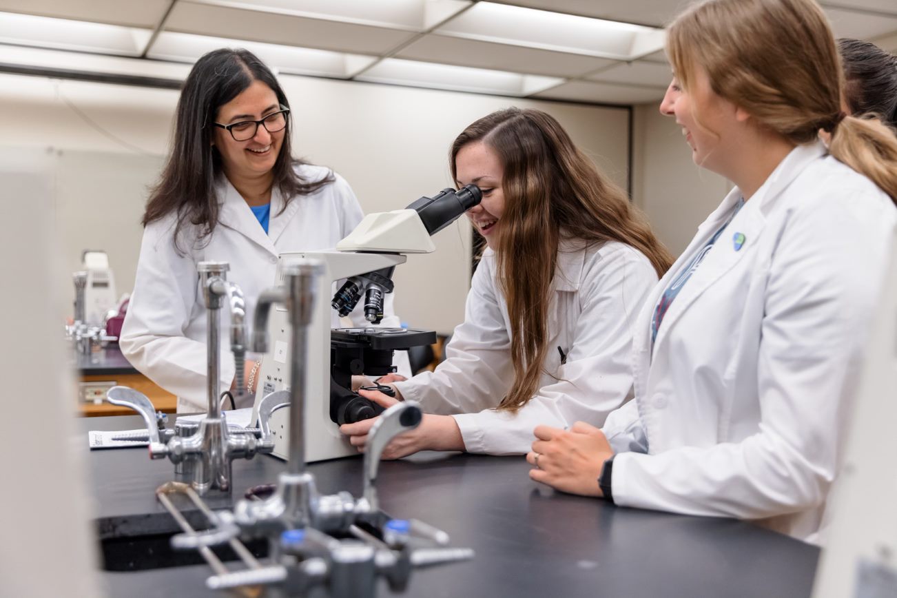 Students in lab coats look through a microscope.