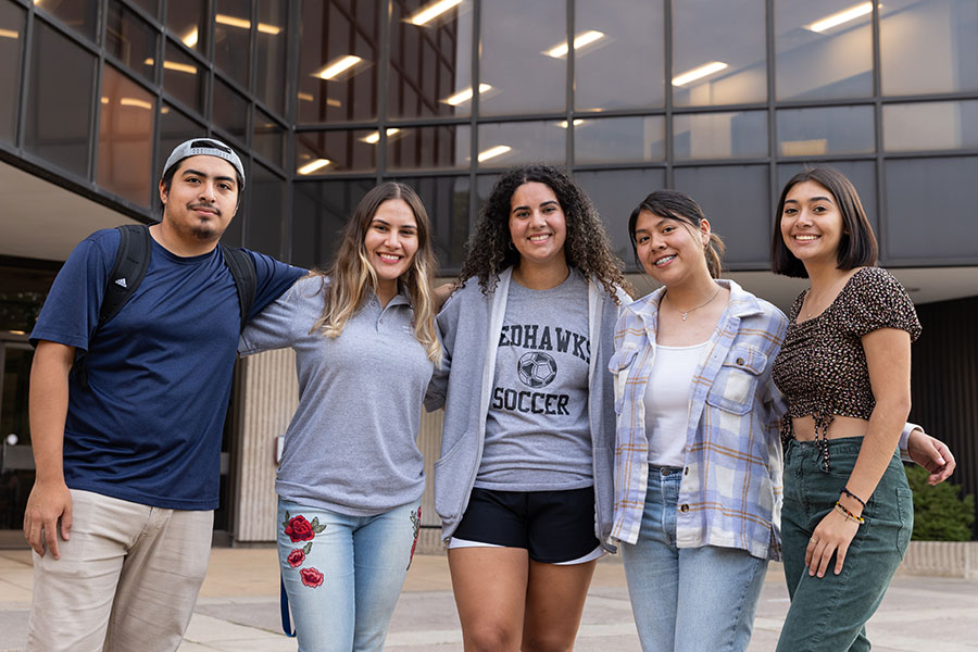 A group of Latino students stands with arms around each other in front of a building.