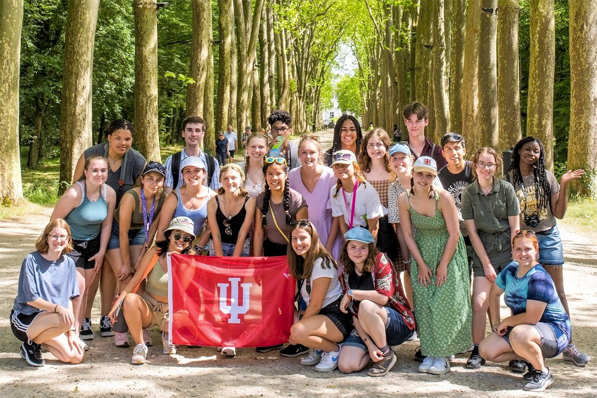A big group of diverse students holding an IU flag in the middle of a tree-lined road.